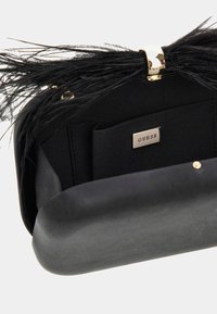 Juoda rankinė-kreminė su plunksnų akcentais, auksinėmis detalėmis ir kontrastinės medžiagos vidumi; turi sidabrinį „GUESS“ logotipą ir kortelių kišenes.