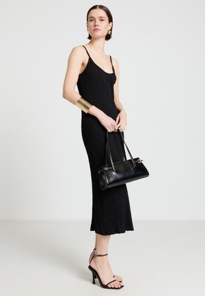 Donna con vestito midi nero senza maniche dal tessuto strutturato e tacchi, che tiene una borsa a mano in pelle nera e indossa un bracciale a polsino e orecchini dorati.