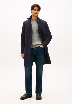 Junger Mann trägt einen dunkelblauen Mantel über einem grauen Pullover und blauen Jeans, steht mit einer Hand in der Tasche, neutraler Hintergrund.