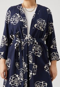 Marinblå morgonrock med vitt blommönster, med bred krage, skärp i midjan och långa ärmar. Tillverkad av ett lätt tyg med synlig textur.