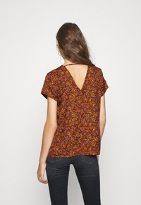 Top marrón con estampado floral, de mangas cortas y diseño en V en la espalda, confeccionado en tejido ligero, combinado con jeans oscuros.