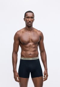 Male model with muscular build wearing black Björn Borg boxer briefs with beige branded waistband against plain white background.