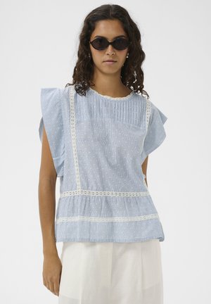 Göndör hajú nő fekete napszemüvegben, világoskék blúzban, fehér csipkedíszítéssel és fehér nadrágban, egy egyszínű háttér előtt állva.