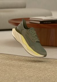 Αθλητικό παπούτσι Clarks σε ελαιί πράσινο με κρέμ και κίτρινη σόλα, αιωρούμενο πάνω από ένα υφασμένο μπεζ χαλί, ξύλινο στρογγυλό τραπέζι και καναπέ στο φόντο.