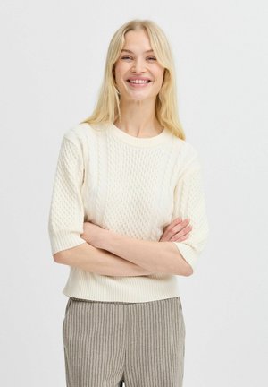 Cremfarbener gestrickter Pullover mit einem strukturierten Zopfmuster, kurzen Ärmeln und geripptem Saum. Getragen mit grauen Nadelstreifenhosen.
