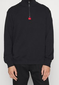 Sort pulloverjakke lavet af blødt stof, med høj krave, halv lynlås foran og en lille rød logoaccent over lynlåsen.