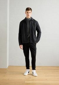 Svart skjortejakke over en grå genser, kombinert med svarte cargo-bukser og hvite joggesko. Jevn tekstur, uformell design.