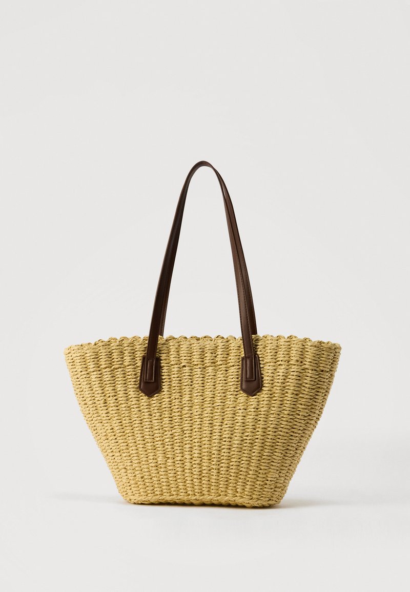 Sac cabas en paille tissée avec deux longues anses en cuir brun foncé, large en haut, étroit à la base, sur un fond uni clair.