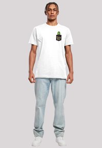 Hvit bomullst-skjorte med et grønt pickle-motiv på en svart lomme med gul tekst. Løs passform, parret med lyseblå jeans og hvite joggesko.