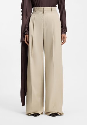 Beige viddebukser med frontplisseringer båret med sorte spidse sko og en mørkebrun top med lange ærmer, stående mod en hvid baggrund.