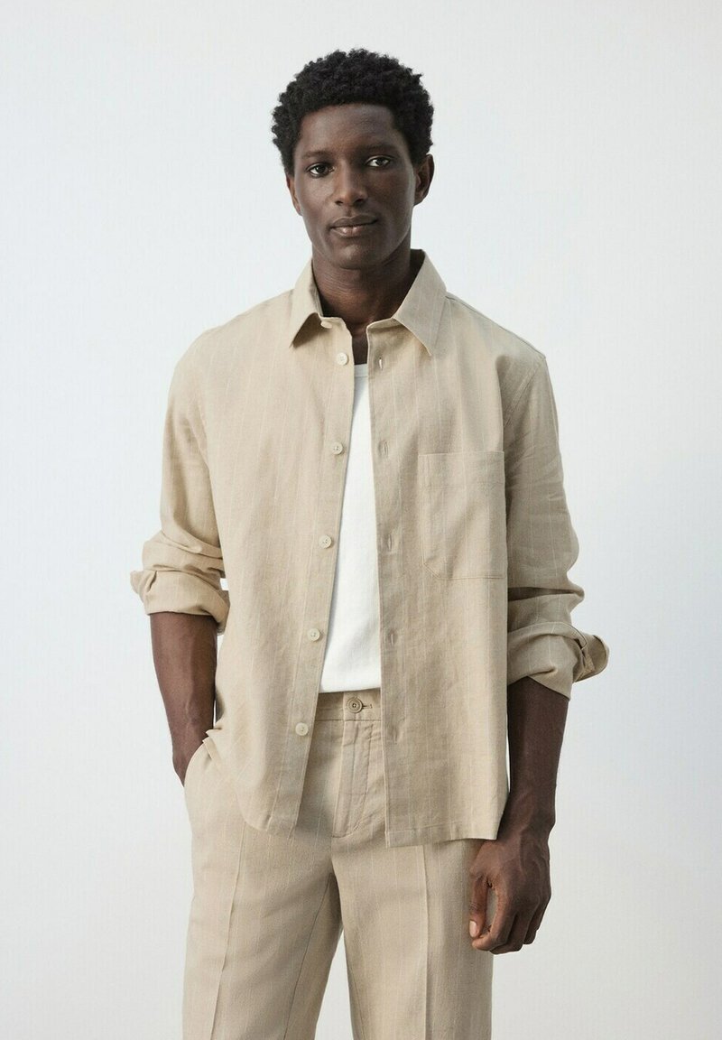 Homme portant une chemise beige à boutons sur un t-shirt blanc et un pantalon beige assorti, debout les mains dans les poches devant un fond uni.