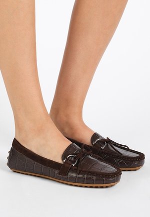 Brune loafers i krokodillemønstret læder med ruskindsdetaljer og kvastudsmykninger, båret på bare fødder mod en hvid baggrund.