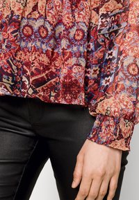 Blusa colorata con motivo floreale e geometrico in rosso, blu e crema. Tessuto leggero con polsini arricciati e maniche lunghe.