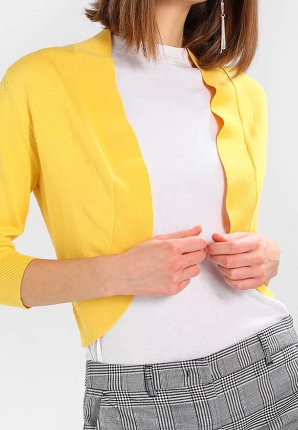 Gele cardigan met driekwart mouwen en een geschaafd voorpand, gedragen over een wit shirt. Gecombineerd met grijze geruite broek.