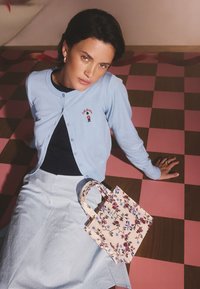 Lyseblå cardigan med knaplukning og broderet detalje, sat sammen med en navyblå top og lyseblå nederdel, ledsaget af en blomsterprintet mulepose.