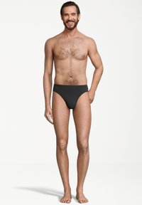 Glunderende man met een baard en borsthaar, gekleed in zwart tekening briefondergoed, staand op blote voeten tegen een witte achtergrond.