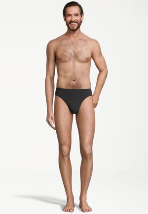 Glunderende man met een baard en borsthaar, gekleed in zwart tekening briefondergoed, staand op blote voeten tegen een witte achtergrond.