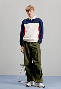 Felpa FILA con pannelli blu navy, bianchi e rossi; pantaloni cargo larghi color olive; scarpe da ginnastica bianche; sfondo grigio semplice.