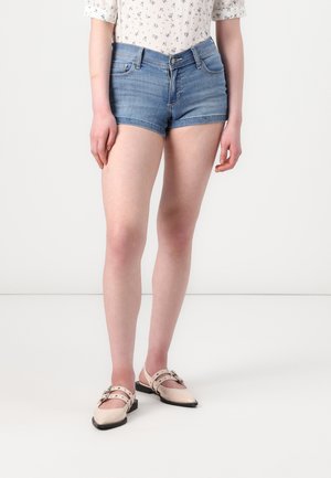 Persona que lleva pantalones cortos de mezclilla azul claro, una camisa blanca con flores y mangas remangadas, y zapatos planos beige con hebillas.