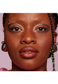 Primo piano di un volto con pelle scura. Tra gli elementi in evidenza ci sono un trucco occhi nero audace, labbra marroni, orecchini grandi e capelli intrecciati con accenti verdi.