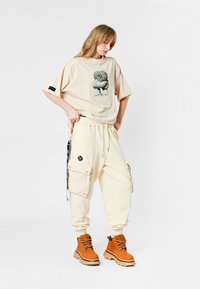 Camiseta oversized beige con gráfico, pantalones cargo sueltos en crema con bolsillos y cremalleras, combinados con botas marrones con cordones. Fondo blanco.
