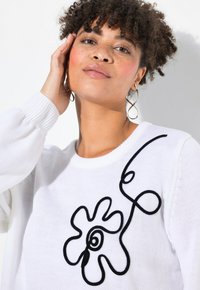 Vitt stickat tröja med en svart blommig broderidesign på framsidan. Ärmarna är ballongstil med ribbade mudd.