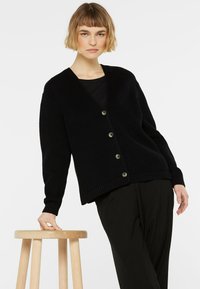 Schwarzer gerippter Cardigan mit V-Ausschnitt, sechsknöpfiger Verschluss und gestrickter Textur. Über einem schwarzen Oberteil getragen und mit schwarzen Hosen kombiniert.