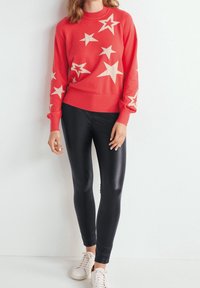 Roter Pullover mit cremefarbenem Sternmuster, geripptem Kragen und Bündchen, kombiniert mit schwarzen Leggings und weißen Turnschuhen vor einem neutralen Hintergrund.