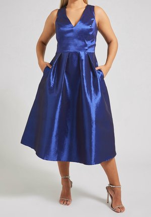 Bez rękawów, błyszcząca chabrowa sukienka za kolano z dekoltem w serek i plisowaną spódnicą, noszona z srebrnymi sandałami na wysokim obcasie, ręce w kieszeniach.