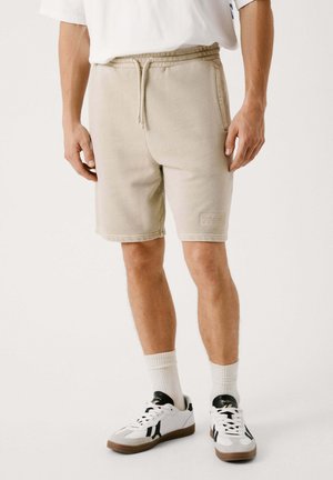 Pantaloni corti in cotone beige con vita elastica e cordino, dotati di tasche laterali e di una piccola etichetta logo sulla gamba sinistra.