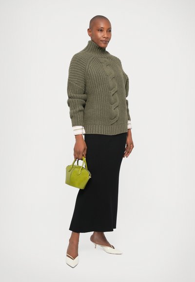 Sweater verde oliva com riscas e colarinho alto, detalhes em ponto de cabos, combinado com uma saia preta e sapatos de salto branco de bico fino, segurando uma pequena carteira verde.