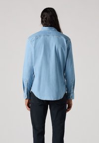 Camicia in denim azzurro chiaro con maniche lunghe, polsini a bottoni e orlo arrotondato. Presenta un colletto classico e dettagli cuciti.