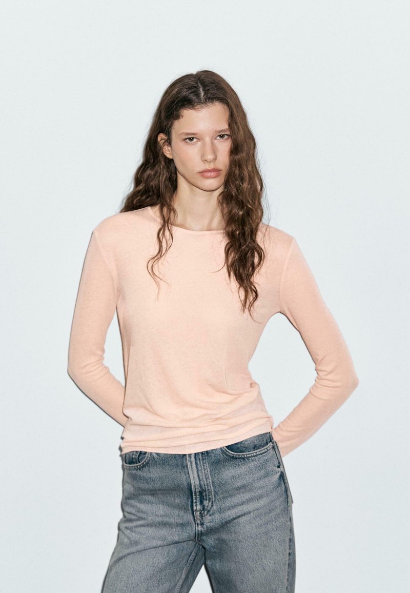 Junge Frau mit langen, welligen Haaren, die ein leicht rosa langärmliges Oberteil und blaue Jeans trägt, steht mit den Händen hinter ihrem Rücken.