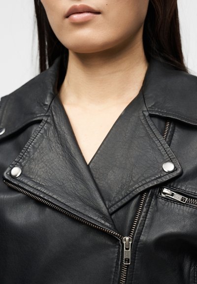 Κοντινή λήψη ενός ατόμου που φορά μαύρο δερμάτινο μπουφάν με ασημένιο φερμουάρ και κουμπιά με σούστα, που δείχνει το γιακά και το πάνω μέρος του στήθους.