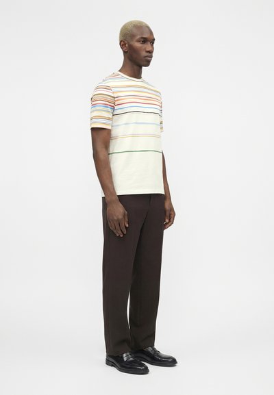 Άνδρας με ξανθά μαλλιά που φοράει ένα κοντομάνικο πολύχρωμο οριζόντιο ριγέ πουκάμισο, σκούρα καφέ παντελόνια και μαύρα επίσημα παπούτσια, στέκεται ακίνητος.