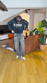 Melna kapuce ar baltu pilienu tekstu, pelēkas platajās bikses ar izbalējušām rakstiem un sudraba sporta apaviem. iekštelpu vide ar augiem un mēbelēm.