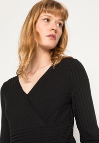 Vestido preto com nervuras, com decote em V e mangas longas, apresentando um tecido texturizado e uma silhueta ajustada.