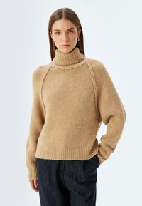Maglione beige lavorato a maglia con colletto alto a costine, maniche raglan e orlo a costine. Texture morbida, vestibilità rilassata e dettagli minimi.