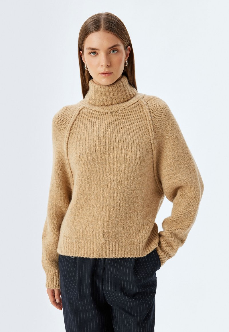 Maglione beige lavorato a maglia con colletto alto a costine, maniche raglan e orlo a costine. Texture morbida, vestibilità rilassata e dettagli minimi.