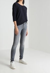 Marinblå tröja med trekvartslånga ärmar, kombinerad med ljusgrå tajta jeans och vita sneakers, på en person som står mot en neutral bakgrund.