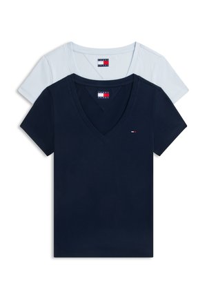 Νεαρή γυναίκα φοράει λευκό T-shirt με V-λαιμόκοψη, μπλε παντελόνια με κάθετες ρίγες, μαύρα παπούτσια και κρατά ένα τζιν μπουφάν στον ώμο της.