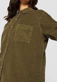 Koszula z oliwkowego sztruksu z zapięciem na guziki, kołnierzykiem, długimi rękawami i kieszonką na piersi, prezentująca fakturowany wzór materiału.