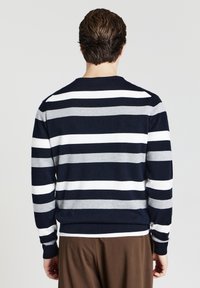 Un homme de dos portant un pull rayé horizontalement bleu marine, blanc et gris avec un pantalon marron, devant un fond uni.