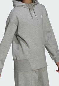 Personne portant un sweat à capuche Adidas gris clair et un pantalon de survêtement assorti, le sweat à capuche arbore le logo Adidas sur la poitrine ainsi que des cordons à la capuche.