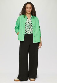 Hellgrüne gesteppte Jacke mit Knöpfen vorne, kombiniert mit einer blusenähnlichen Oberbekleidung mit geometrischem Muster und weitem schwarzen Hosen. Lässige Schuhe runden den Look ab.