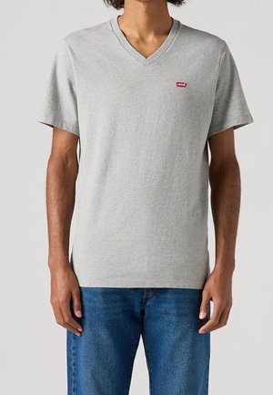 Mies, jolla on päällään vaaleanharmaa V-kaula-aukollinen T-paita, jossa on pieni punainen Levi's-logo rinnassa, ja siniset farkut, seisoo yksinkertaisen taustan edessä.