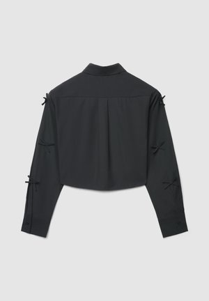 Schwarze, langärmlige, bauchfreie Bluse mit kleinen gebundenen Schleifen an den Ärmeln, geradem Kragen, Rückenfalte und knöpfbaren Manschetten vor grauem Hintergrund.