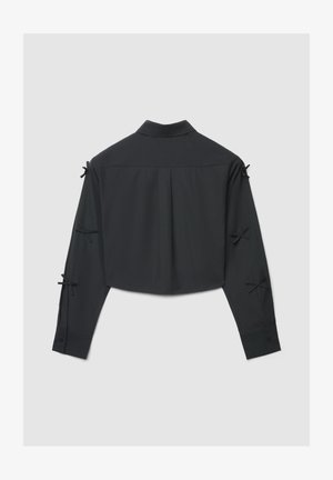 Schwarze, langärmlige, bauchfreie Bluse mit kleinen gebundenen Schleifen an den Ärmeln, geradem Kragen, Rückenfalte und knöpfbaren Manschetten vor grauem Hintergrund.