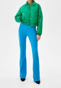Zelená prošívaná zkrácená bunda s patentkami; v kombinaci s přiléhavými jasně modrými rozšířenými kalhotami a černými špičatými balerínami.