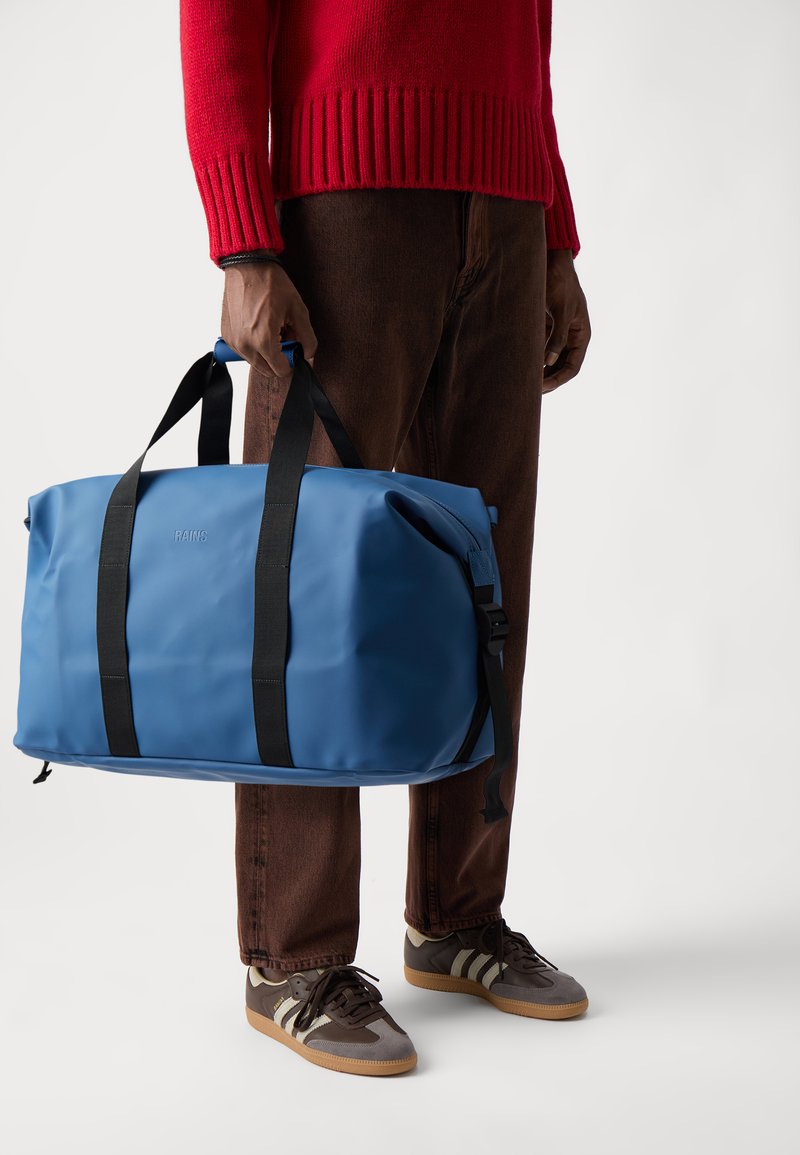 Sac de sport étanche bleu avec des poignées en toile noires et une poche zippée sur le côté, fabriqué à partir d'un matériau lisse et durable, tenu par une personne.